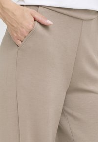 Gai bēša bikses, kas izgatavotas no maiga, elastīga auduma, ar sānu kabatu, kas izceļas ar elegantu dizainu un patīkamu tekstūru.