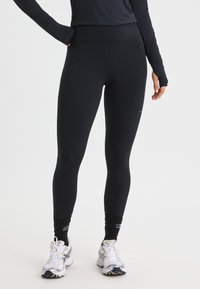 Black stretchy athletic leggings with a high waist, smooth texture, and minimal branding, paired with white running shoes and black socks.