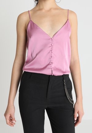 Vrouw draagt een roze satijnen hemdje met knopen aan de voorkant en zwarte broek met een zilveren kettingaccessoire aan de rechterkant.