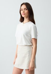 T-shirt en coton blanc à manches courtes, associé à une mini jupe en lin beige clair. Un collier en or ajoute un contraste subtil. Design simple et décontracté.