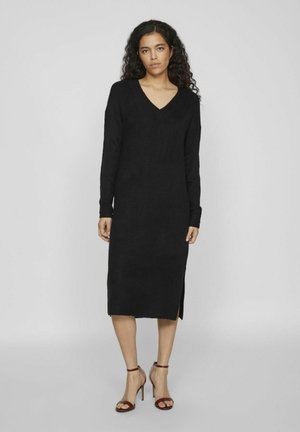 Vestido negro de punto con mangas largas y escote en V, que presenta una abertura lateral. La tela parece suave. Combinado con sandalias de tacón abierto.