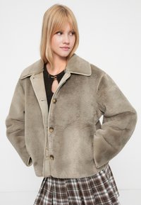 Veste en fausse fourrure beige avec un col classique, des boutons à l'avant et des poches latérales, arborant une texture douce et une silhouette décontractée.
