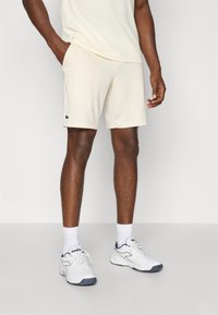 Pantaloni sportivi beige chiaro in un tessuto morbido, con tasche laterali e un piccolo logo verde scuro. Abbinati a calzini e scarpe bianche.