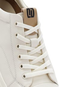 Baskets en cuir blanc avec des lacets plats blancs. Présente un accent en cuir beige sur le talon avec un logo noir. Texture lisse et design minimaliste.