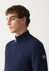 Ung mand med krøllet hår iført en mørkeblå striktrøje med halv lynlås og Colmar-logo på venstre ærme.