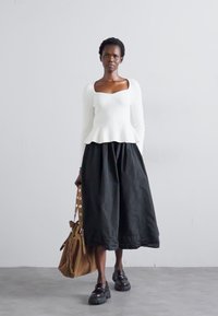 Weißes, geripptes Langarmoberteil mit peplum Saum, kombiniert mit einem schwarzen, fließenden, knöchellangen Rock und einer braunen Schultertasche. Klobige schwarze Schuhe.
