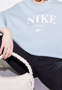 Světlomodrá mikina s bílým logem "NIKE". Černé kalhoty, drží pletenou bílou tašku. Hladká textura, volný styl.