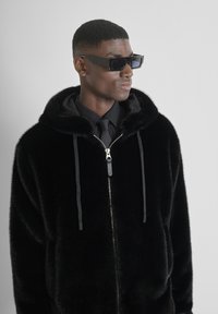 Crna jakna s kapuljačom od umjetnog krzna, s patentnim zatvaračem, detaljima na vezice i glatkom satenskom unutrašnjom podstavom. Uparena sa elegantnim crnim sunčanim naočalama.