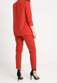 Czerwona marynarka i dopasowane spodnie, wykonane z gładkiej tkaniny. Marynarka ma kołnierz i podwinięte rękawy, a spodnie mają zwężany krój. Czarne sandały na obcasie.