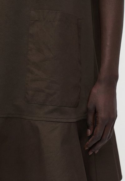 Χέρι με σκούρο δέρμα δίπλα σε καφέ ένδυμα με μεγάλη τετράγωνη τσέπη στο μπροστινό μέρος, που δείχνει την υφή του υφάσματος και τις λεπτομέρειες της ραφής.