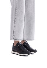 Zapatillas atléticas negras con acentos de malla y cuero, que presentan un diseño elegante, punta redonda y suela blanca contrastante. Combinadas con jeans de pierna ancha gris.