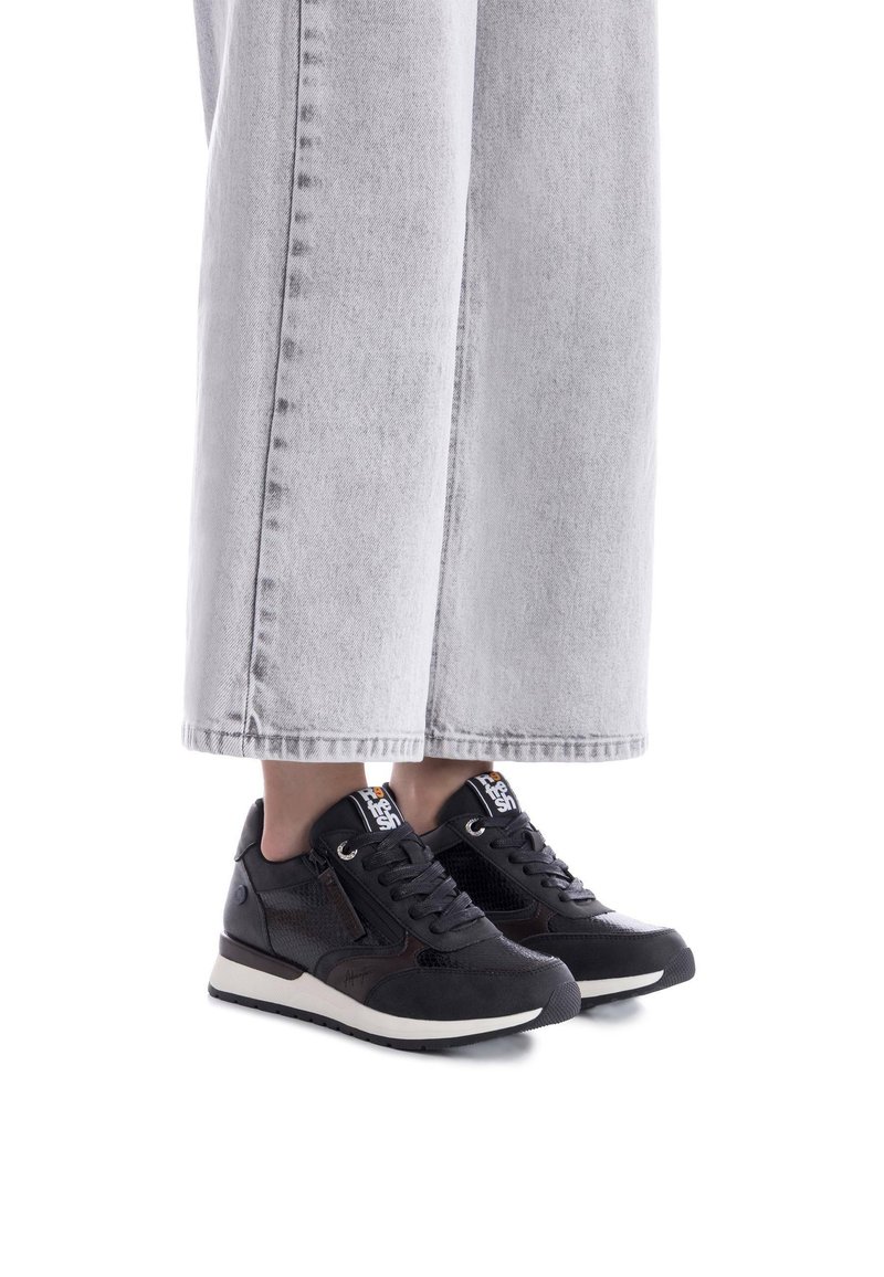Zapatillas atléticas negras con acentos de malla y cuero, que presentan un diseño elegante, punta redonda y suela blanca contrastante. Combinadas con jeans de pierna ancha gris.