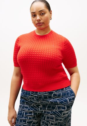 Žena u crvenom kratkom rukavu džempera s kabelskim uzorkom i mornarskoplavim uzorkovanim hlačama, ruke u džepovima, na bijeloj pozadini.