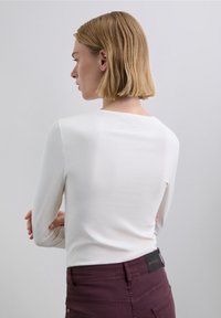 Top blanco de manga larga confeccionado en una tela suave, con escote redondo; combinado con pantalones ajustados de color granate oscuro que muestran un detalle de bolsillo trasero.