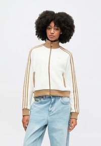 Chaqueta blanca corta con cremallera, con puños y cuello acanalados marrones, que cuenta con tres rayas marrones en cada manga y dos bolsillos laterales.
