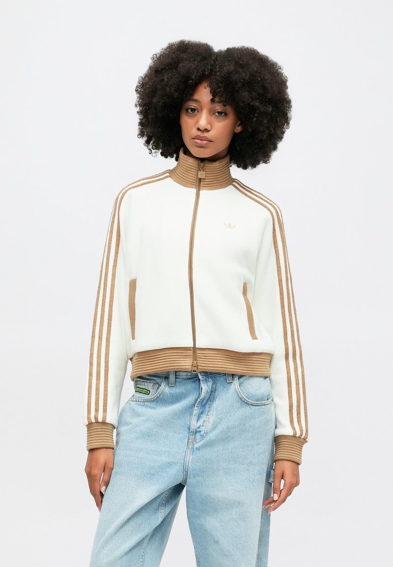 Veste blanche zippée coupée avec des poignets et un col côtelé marron, comportant trois bandes marron sur chaque manche et deux poches latérales.