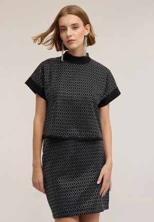 Młoda kobieta z krótkimi włosami, ubrana w czarno-biały top w romby z krótkim rękawem i pasującą spódnicę, długie srebrne kolczyki z frędzlami.