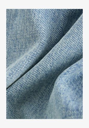 Tissu en denim bleu clair présentant une texture lisse, un motif de tissage diagonal et des plis doux, montrant des variations subtiles de couleur.