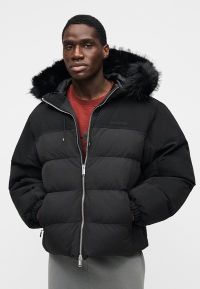 Casaco acolchoado preto com capuz de pelo falso, fecho de correr e punhos elásticos. Feito de tecido texturizado com padrões acolchoados horizontais.