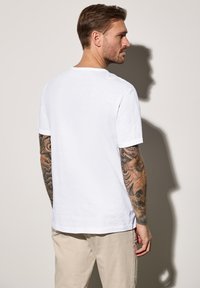 Bílý krátký rukáv tričko s kulatým výstřihem a texturovaným povrchem. Na rukávu viditelné tetování a světlé kalhoty.