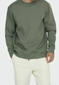 Felpa verde oliva con scollo a girocollo e polsini a costine. Tessuto morbido, vestibilità comoda, abbinata a pantaloni in velluto a coste di colore chiaro.