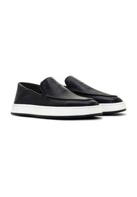 Black leather slip-on shoes with textured upper, white rubber sole, and stitch detailing. Rounded toe and minimal design.