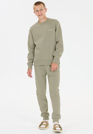 Adolescent debout, portant un sweat-shirt beige et un pantalon assorti, des chaussettes blanches et des sandales beiges, sur un fond blanc uni.