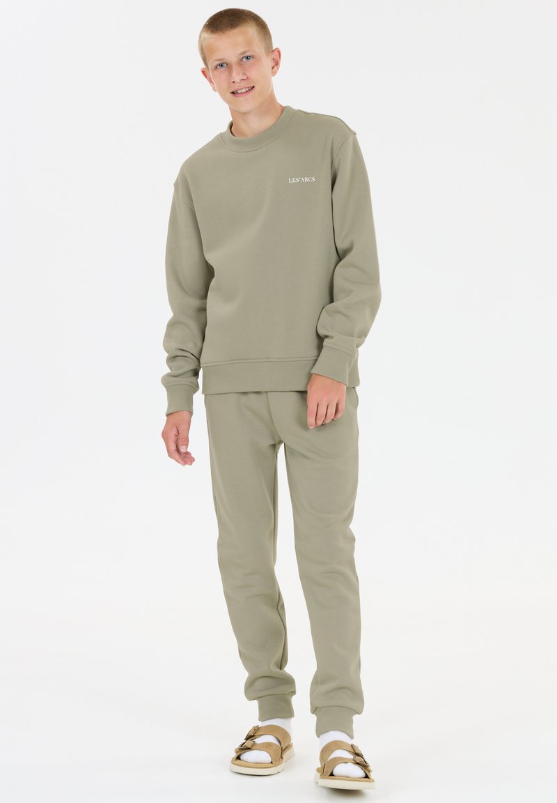 Adolescent debout, portant un sweat-shirt beige et un pantalon assorti, des chaussettes blanches et des sandales beiges, sur un fond blanc uni.