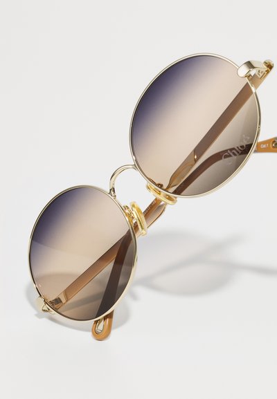 Gafas de sol redondas con marco de metal dorado, lentes marrones degradados y varillas beige. Diseño notable de doble puente y logotipo en la lente.