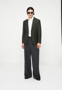 Dunkelgrüner Blazer, weißes Hemd und weite graue Hose. Glänzende schwarze Schuhe, elegante Sonnenbrille. Glattes Material, moderne Silhouette, minimalistisches Design.
