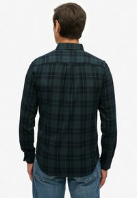 Man die met de rug naar ons staat, gekleed in een donkergroene en blauwe geruite lange mouwen shirt en blauwe jeans, staande tegen een witte achtergrond.