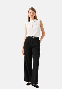 Frau trägt eine ärmellose weiße Bluse, schwarze hochtaillierte weite Hosen mit Gürtel und schwarze Ballerinas und steht mit einer Hand in der Tasche.