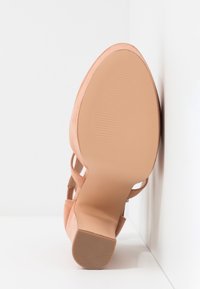 Sandale à talon haut en daim beige, vue semelle vers le haut contre un mur blanc en angle, avec un talon carré et une bride à la cheville.