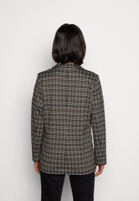 Osoba s krátkými tmavými vlasy, oblékající hnědou, černou a bílou kostkovanou blazer, stojí čelem k jednoduchému bílému pozadí.