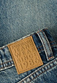 Traperice s svijetloplavim pranjem, s smeškim kožnim etiketom na kojoj je utisnuto "FORWARD JEANS WEAR SINCE 1986".