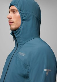 Türkisfarbene Kapuzenjacke aus glattem, wasserabweisendem Material. Mit hohem Kragen, Reißverschluss und Logo auf der Brust und dem Ärmel.