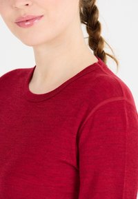 Kvinna med löst flätat hår som bär en röd långärmad tröja, som visar hennes underansikte och axel mot en vit bakgrund.