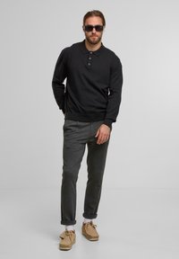 Mann in schwarzem Langarm-Polohemd, grauen Hosen, hellbraunen Wildlederschuhen, weißen Socken und schwarzen Sonnenbrillen, der vor einem schlichten Hintergrund steht.