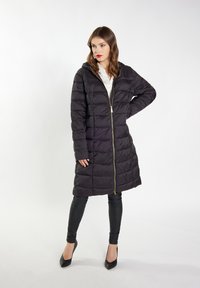 Zwarte pufferjas met capuchon, voorzien van een gouden ritssluiting. Verticaal gelegde quiltpatroon en halflang. Gekocht over zwarte leggings en hakken.