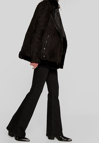 Chaqueta de pelo sintético negra con un alto cuello, cierre de cremallera en la parte delantera y bolsillos laterales, combinada con jeans de campana negros y botas de tobillo negras brillantes.