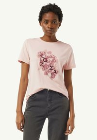 Light pink cotton t-shirt with short sleeves featuring a graphic print in dark pink and brown abstract shapes, centered text in black.