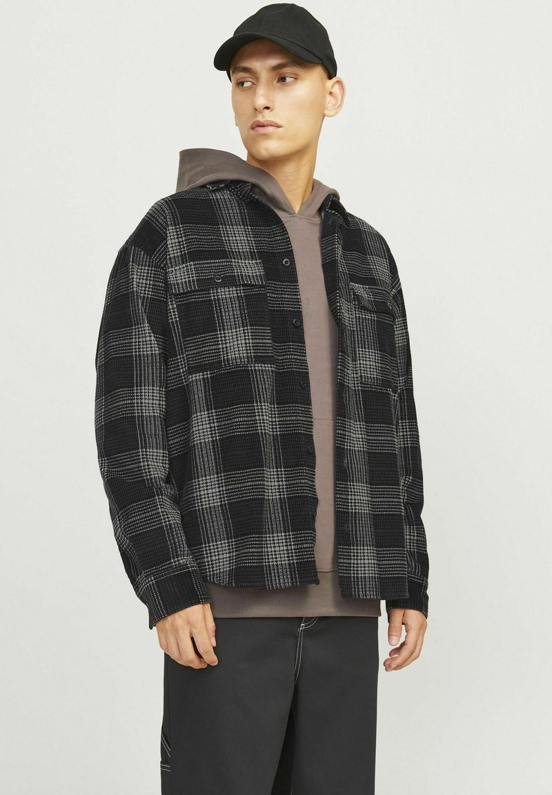 Zwart en grijs geblokt flanellen shirt met twee borstzakken, gelaagd over een bruine hoodie. Model draagt een zwarte pet en donkere broek.