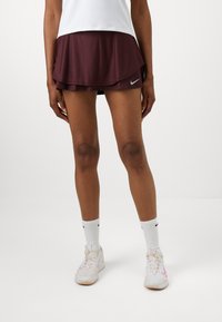 Burgundy sport szoknya réteges kivitelben, fehér crew zoknikkal és rózsaszín díszítésű tornacipőkkel párosítva. Simaság a tapintásban, a Nike logó látható.