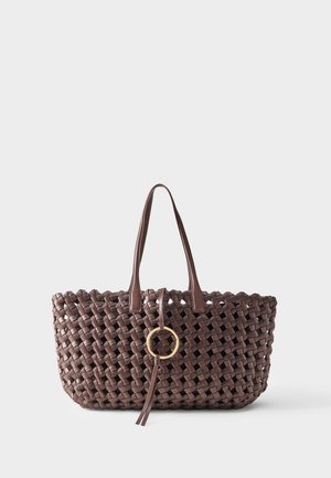 Donkerbruine geweven tote tas met twee handvatten en een decoratieve gouden ring met leren tassels middenvoor.