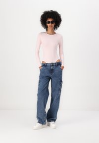 Svetlo roza bodysuit s dolgimi rokavi in izrezi, v kombinaciji z ohlapnimi modrimi cargo hlačami z bočnimi žepi in belimi športnimi čevlji; elegantna črna sončna očala.