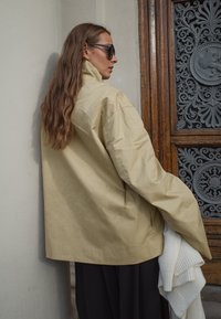 Prevelik bež plašč iz lahkega materiala, z visokim ovratnikom in sproščenim krojem, v kombinaciji z kremno barvo pletenim ovitkom.