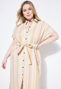 Abito camicia a righe in crema con strisce pastello giallo, viola e arancione. Presenta un'apertura con bottoni, maniche corte e una vita con cintura.