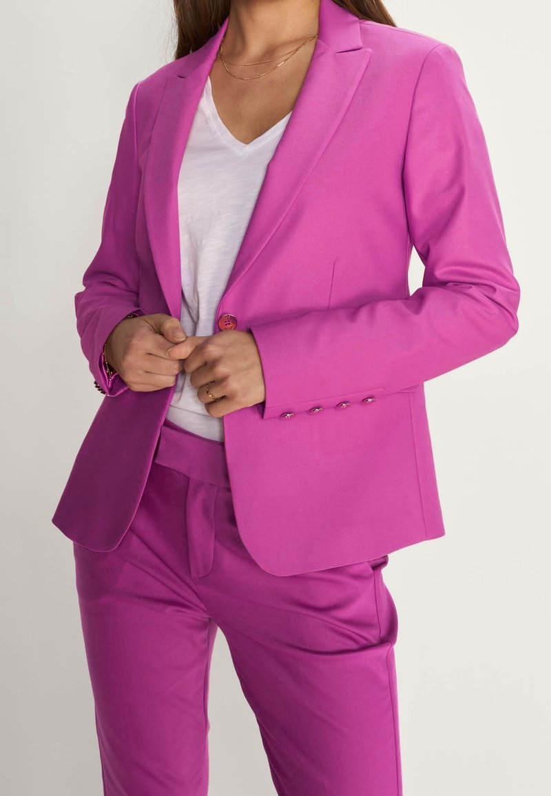 Blazer rose ajusté en tissu lisse, avec une fermeture à un bouton, des revers crantés et des détails de couture subtils, accompagné d'un pantalon assorti.
