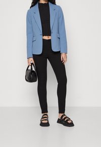 Svetle modré blejzr přes černý krátký top, spárováno s černými úzkými džíny a masivními černými sandály. Drží malou černou kabelku.