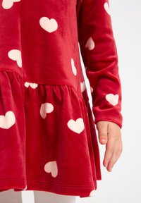 Robe en velours rouge avec des manches longues, dotée d'un ourlet froncé et de motifs de cœurs blancs. Texture douce, coupe ajustée et poignets serrés.